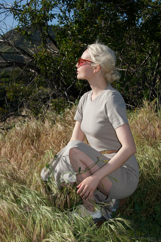 A woman with short blonde hair, wearing sunglasses, kneeling in a field of tall grass and wildflowers. She wears the AWAN T Dress in Beige, blending seamlessly with the natural surroundings.