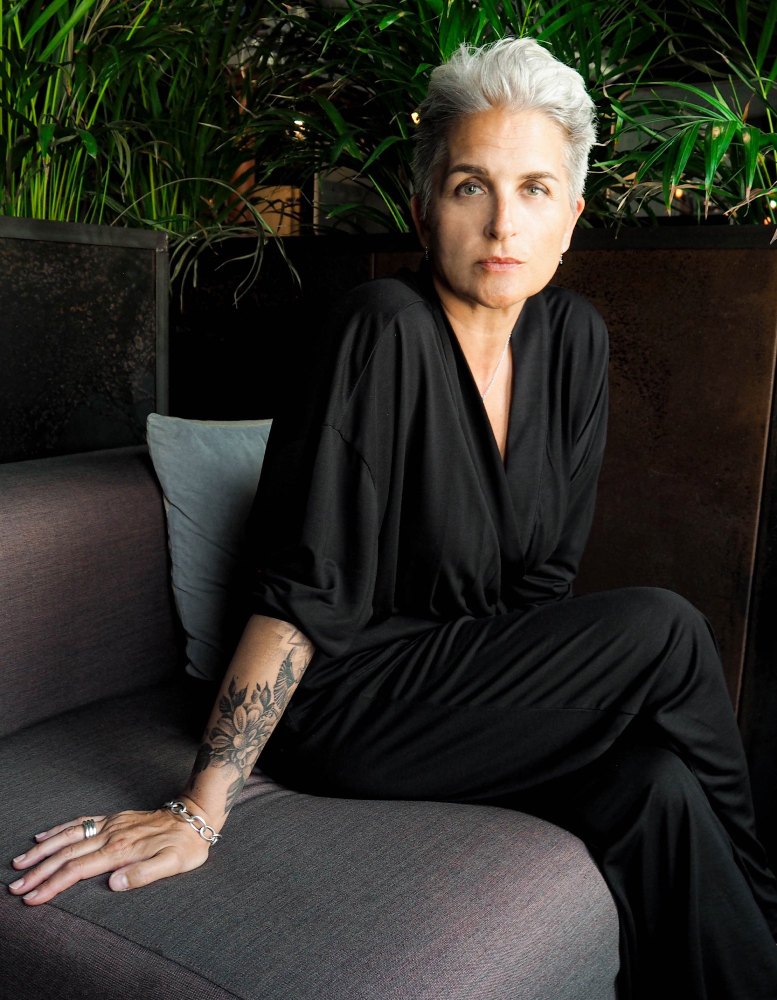 A woman with short silver hair wearing the AWAN Wrap Cardigan in Warm Black, sitting on a sofa in a cozy setting with lush green plants in the background.