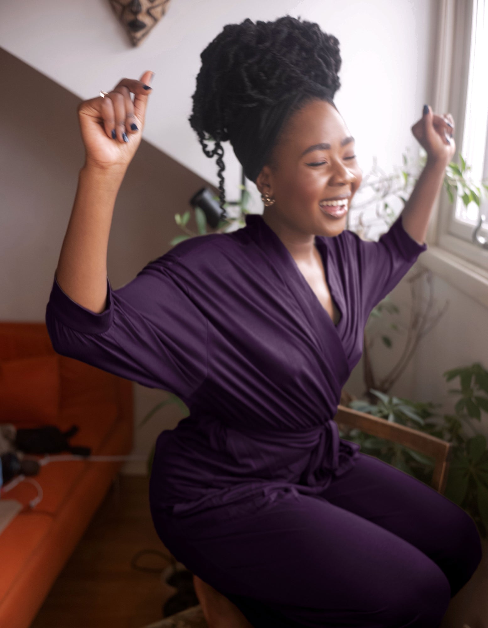 A woman with an afro hairstyle wearing the AWAN Wrap Cardigan in Deep Plum, smiling and dancing joyfully in a bright room with an orange sofa in the background."