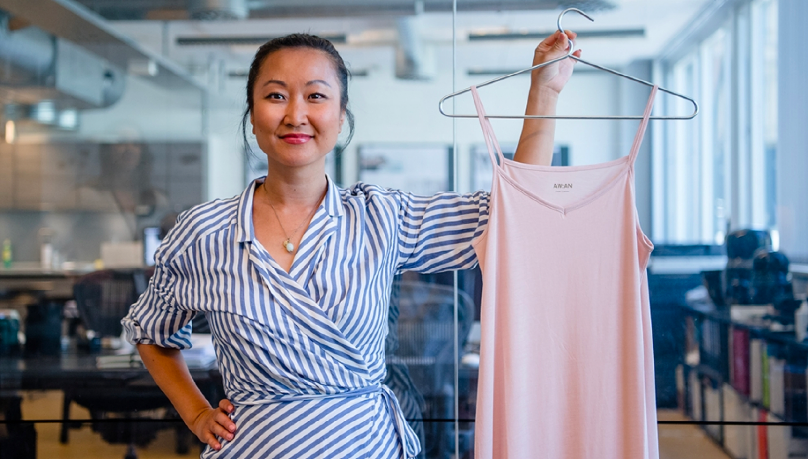 Anette Miwa Dimmen, founder of AWAN, holding up a light pink dress in a modern office setting. She is wearing a blue and white striped dress, smiling confidently. Quote: 'We believe what we choose to wear is just as important as the products we use on our skin.' – Anette Miwa Dimmen, Founder of AWAN. This image reflects AWAN's commitment to high-quality, skin-friendly fashion choices.