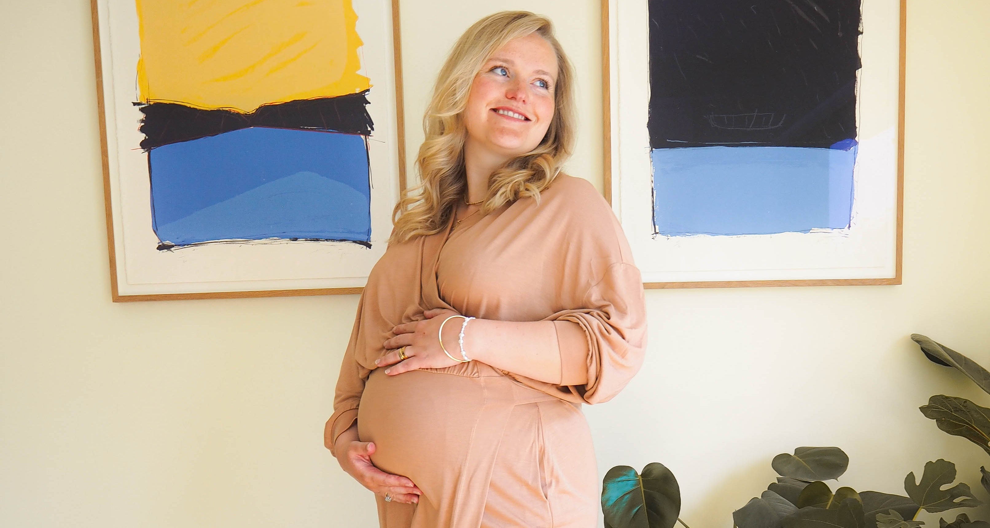 A model stands gracefully in the AWAN Wrap Dress in Camel Rose, posing in front of abstract art pieces with a happy smile and cradling her baby bump.