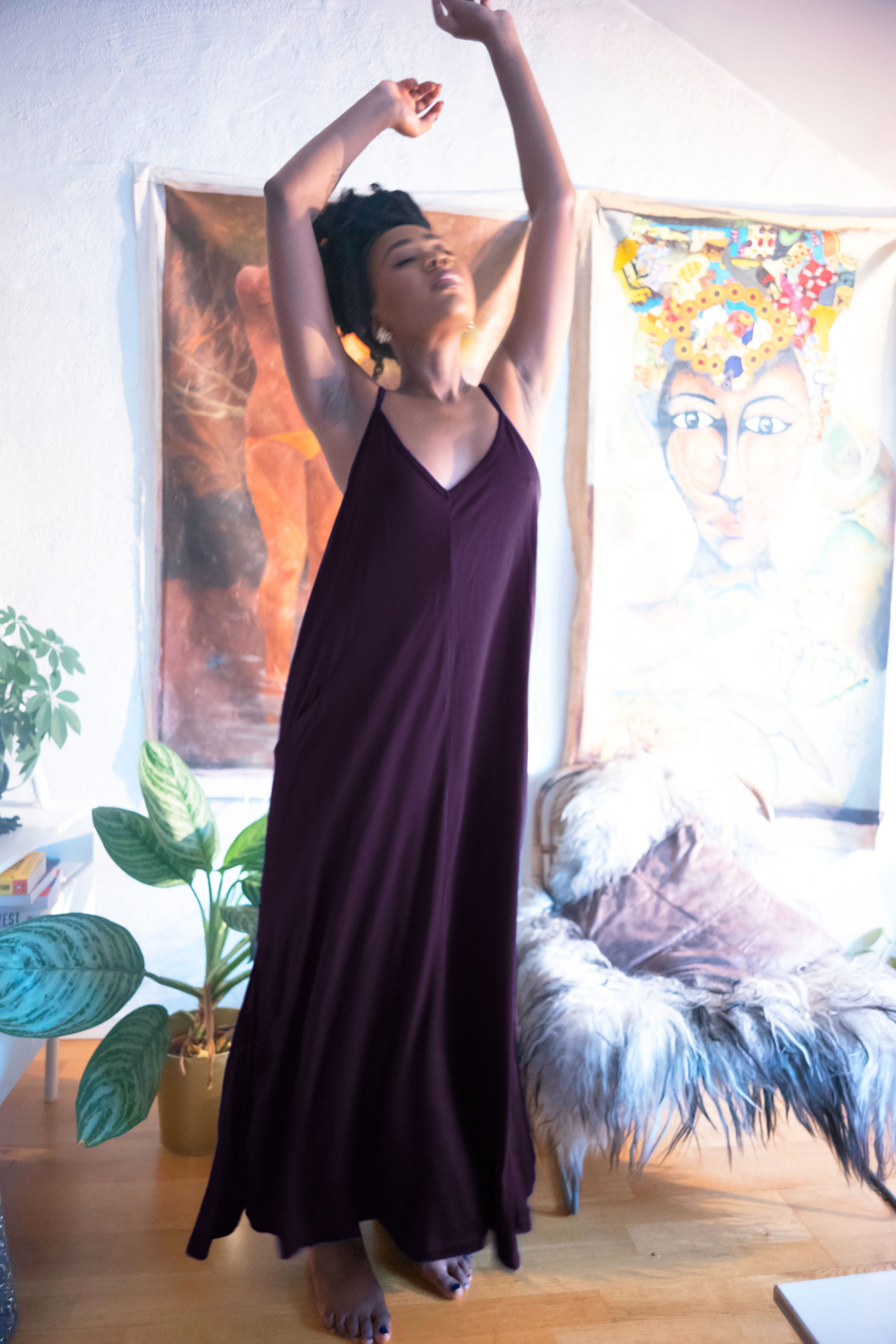 A woman wearing the AWAN Boho Slip Dress in Deep Plum, sitting elegantly on a chair in a modern interior with a potted plant, accessorized with gold jewelry